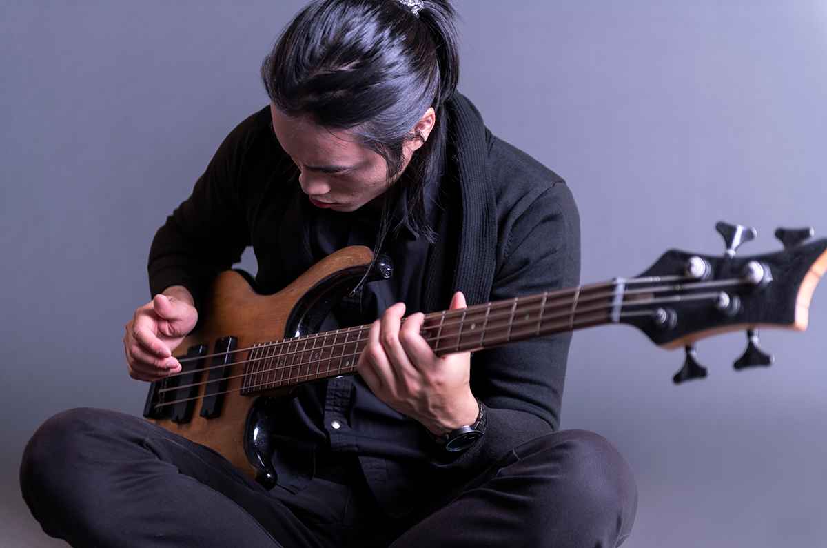 Pria berbaju hitam sedang duduk sambil memainkan bass elektrik berwarna merah.