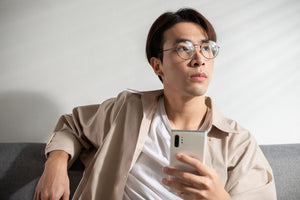 Pria Asia dengan kacamata sedang berpikir sambil memegang ponsel.